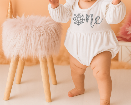 Baby girl in a purple ‘Onederland’ snowflake outfit, standing beside a fluffy stool on a soft lavender background — perfect for a snowflake Onederland 1st birthday cake smash photoshoot.