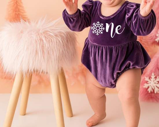 Baby girl in a purple ‘Onederland’ snowflake outfit, standing beside a fluffy stool on a soft lavender background — perfect for a snowflake Onederland 1st birthday cake smash photoshoot.