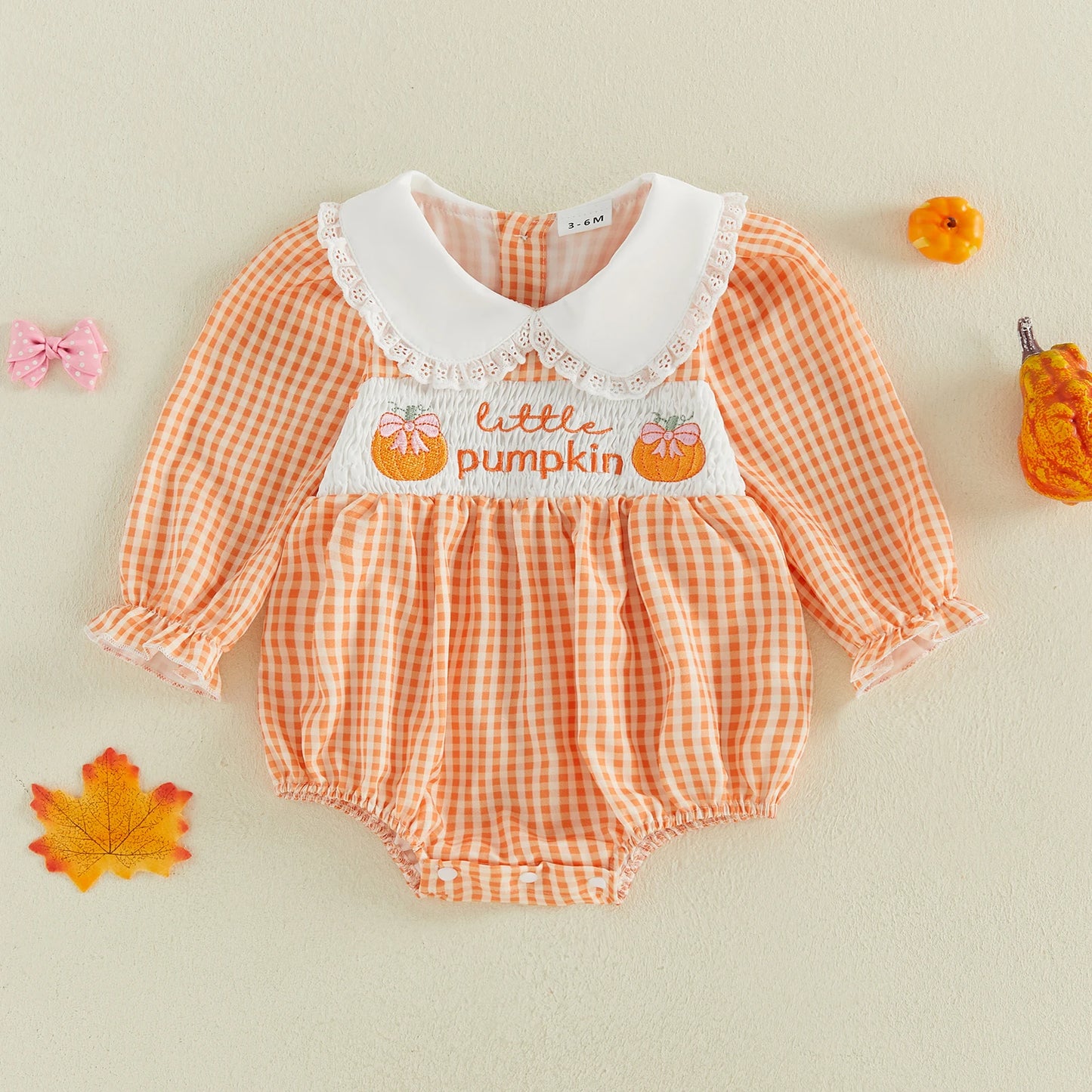 A baby romper with an orange and white gingham pattern, a white lace Peter Pan collar, and 'little pumpkin' embroidered on the chest. The romper is displayed flat on a light beige background with autumn-themed decorative props including a leaf, small pumpkins, and a bow.