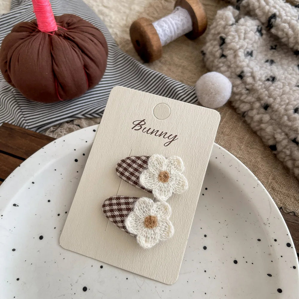 Two small flower hair clips on a cream display card labeled 'Bunny,' styled on a speckled white plate with cozy fabric props in the background.