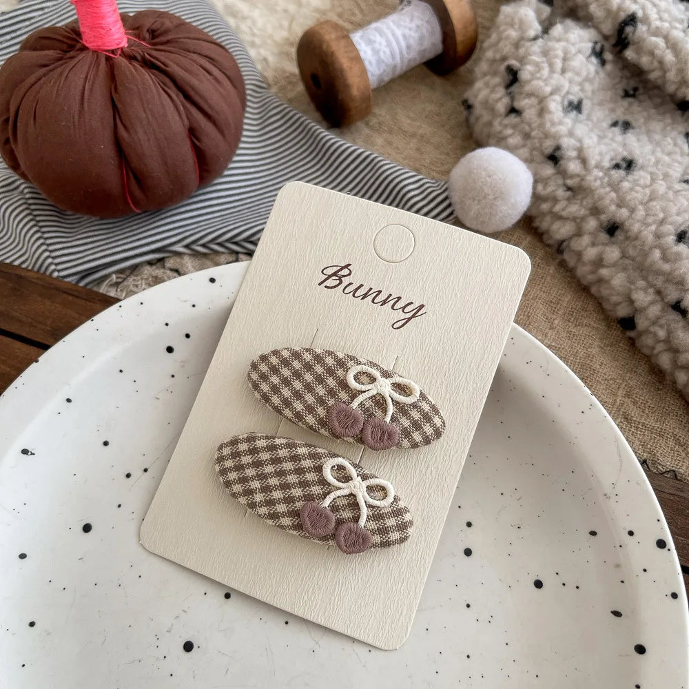 Two small hair claw clips with a brown and beige gingham pattern, white bow details, and pink flower accents, displayed on a cream card labeled 'Bunny'. The card is styled on a speckled white plate with autumn-themed props in the background.