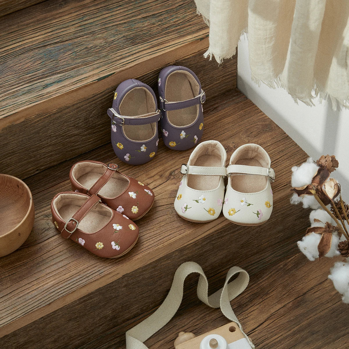 Three pairs of baby Mary Jane shoes arranged on rustic wooden steps: lavender with yellow and white floral embroidery, brown with matching floral embroidery, and cream with small yellow and green floral embroidery. A cream strap and small wooden accessory are visible in the foreground with cotton stems and a wood bowl as styled props.