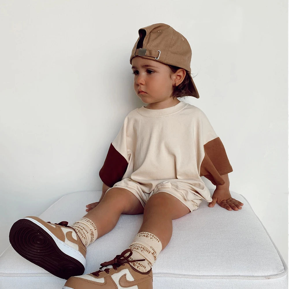 Child wearing a beige cap, white shirt with brown sleeves, beige shorts, and brown shoes sitting on a white surface.