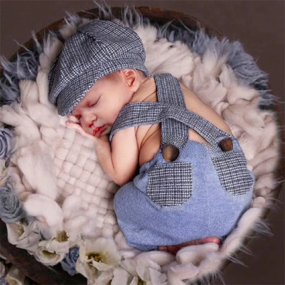 A sleeping newborn baby wearing a matching blue denim-style romper and cap set with a gray-and-white check pattern, posed in a soft white textured blanket basket.