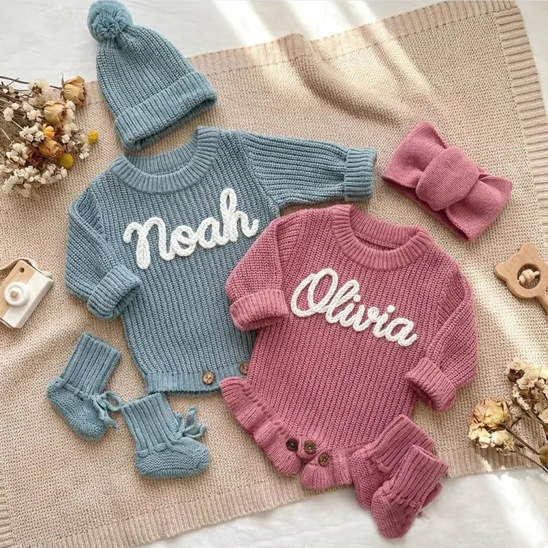 Knitted baby outfit with 'Noah' and 'Olivia' on sweaters, socks, and headband on a textured surface.