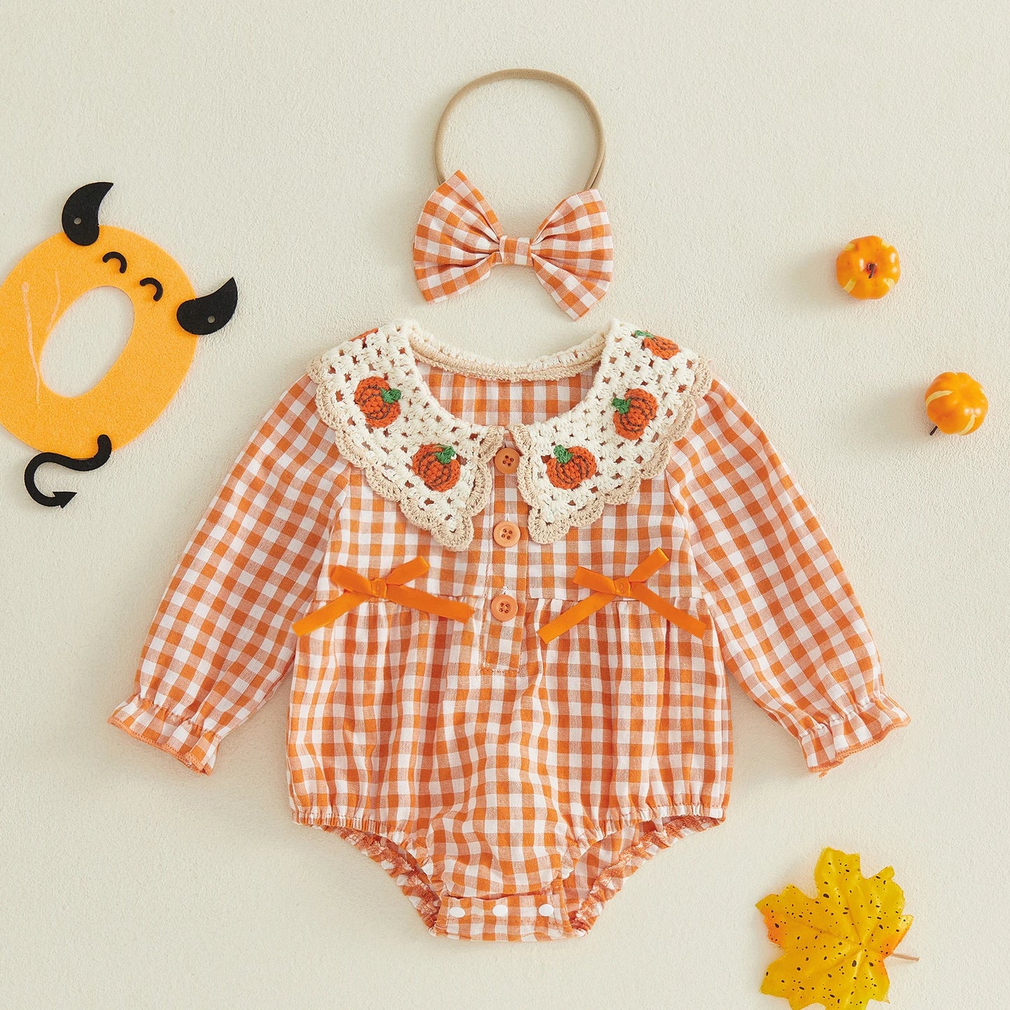 A baby romper set in orange and white gingham, featuring a white crochet Peter Pan collar decorated with small orange pumpkins, orange bow accents on the chest, and a matching orange and white gingham bow headband. The set is styled on a light beige background with autumn-themed props including a yellow pumpkin, a leaf, and small orange pumpkins.