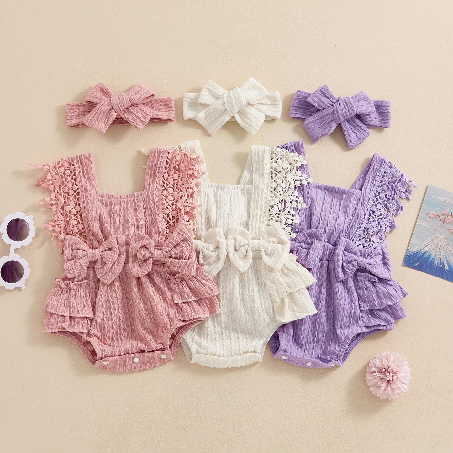 Three baby rompers in pink, white, and purple with matching headbands on a beige background.