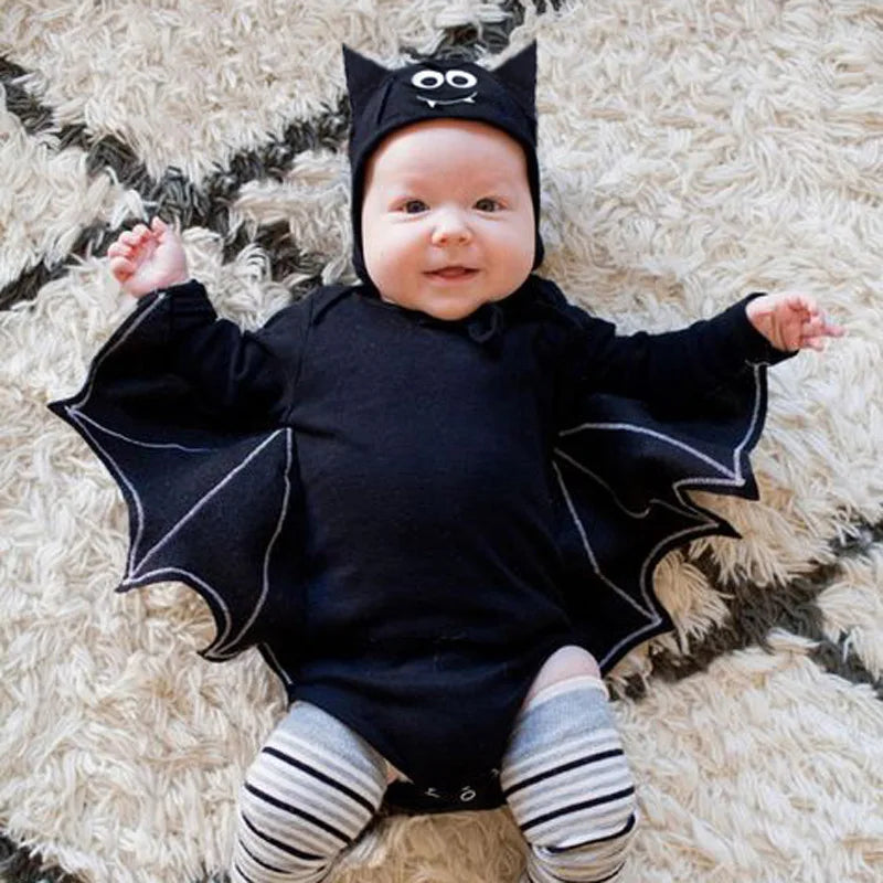 A baby wearing a black bat costume with attached wings, a black hat with bat ears, and striped tights, lying on a shaggy cream rug.