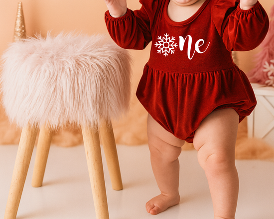 Baby girl in a red ‘Onederland’ snowflake outfit, standing beside a fluffy stool on a soft lavender background — perfect for a snowflake Onederland 1st birthday cake smash photoshoot.