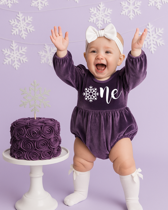 Baby girl in purple Onederland snowflake romper for 1st birthday cake smash photoshoot.
